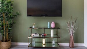 A room with a television mounted on the wall, a glass shelving unit with decorative items, a bamboo plant, and a vase with branches. - A Stylish, essential 3bed Home with Secure Parking (Nottingham)