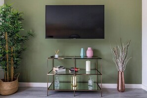 Una habitación con un televisor montado en la pared, un estante de vidrio con objetos decorativos, una planta de bambú y un florero con ramas.