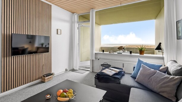 Una sala de estar moderna con techo de madera, un televisor de pantalla plana montado en la pared, un sofá con cojines, una mesa de centro con fruta y una ventana con vistas al cielo.