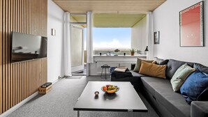 Una sala de estar moderna con un televisor de pantalla plana montado en una pared de paneles de madera, una mesa de centro negra con fruta, un sofá gris con cojines y vistas a la ciudad a través de un ventanal grande.