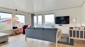 Un salon spacieux avec un canapé gris, un fauteuil rouge, un téléviseur fixé au mur et de grandes fenêtres donnant sur l’extérieur.