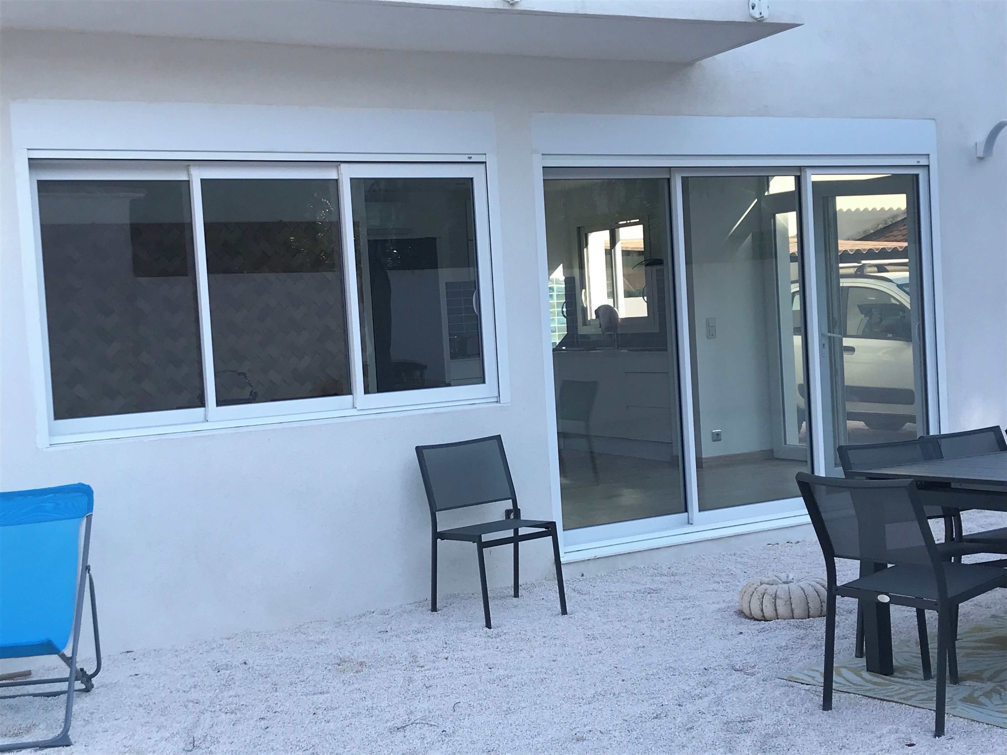 A patio area with a table and chairs, a blue chair, and a large sliding glass door leading to a parking area.