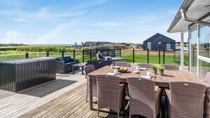 Une terrasse extérieure aménagée avec des meubles en osier et un jacuzzi, offrant une vue sur une pelouse et un plan d’eau.