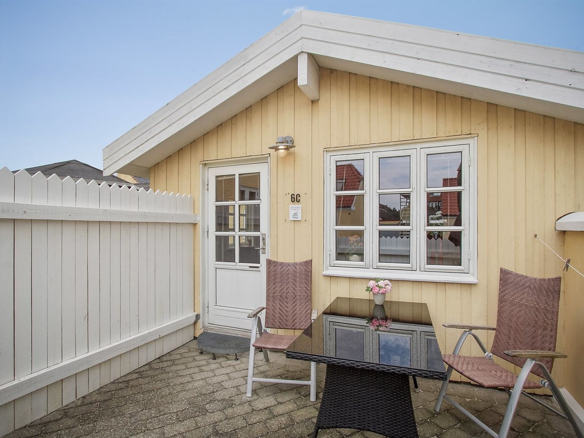 Een klein, laag gebouw met een witte gevel en een houten hek. Er staat een glazen tafel met twee stoelen en een kleed. Een deur met het nummer '6C' is zichtbaar.