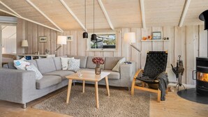 Un salon chaleureux avec un canapé gris, une table basse en bois et un fauteuil noir. On y trouve une cheminée et une fenêtre donnant sur l’extérieur.