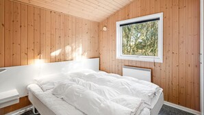 Un dormitorio con paredes de madera, una cama con ropa de cama blanca, una ventana con vistas a árboles y una lámpara en el techo.
