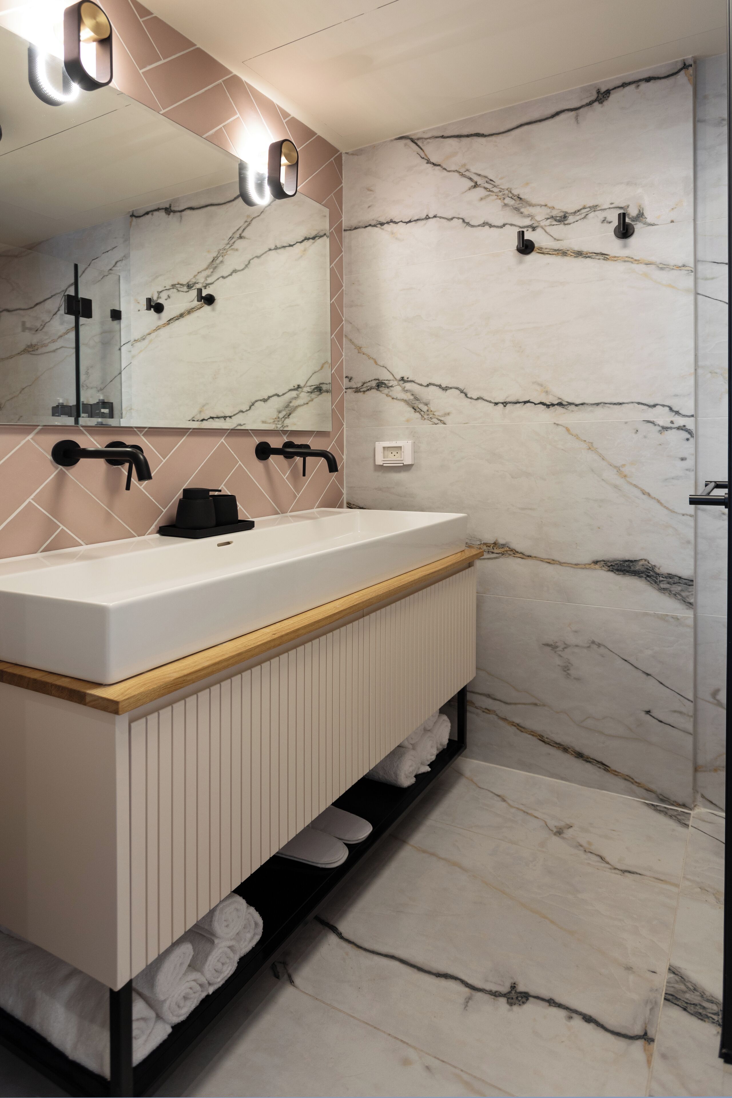 Une salle de bain avec un mur en marbre, un lavabo blanc, des robinets noirs et un meuble-lavabo en bois.