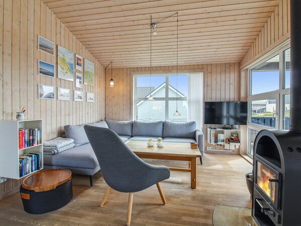A cozy living room with a wooden ceiling, a grey sofa, a wooden coffee table, a black armchair, a bookshelf, a TV, and a fireplace.