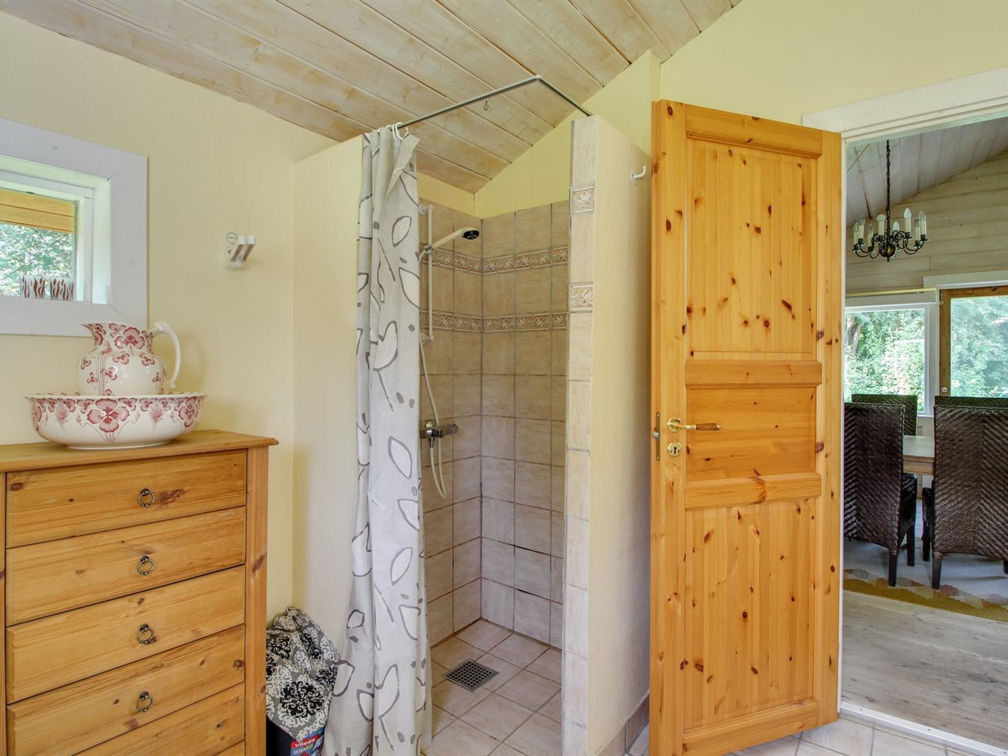 A wooden cabinet with drawers, a shower with tiled walls, and a wooden door leading to a dining area with wicker chairs.