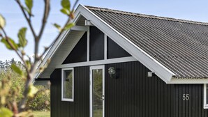 Ein kleines, dunkles Holzgebäude mit weißer Tür und Fenster, Hausnummer 55, eingebettet in eine grüne Wiese mit Bäumen im Hintergrund.