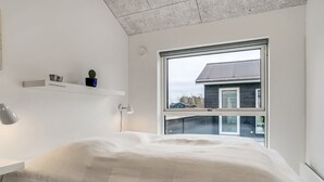 Une chambre moderne avec une grande fenêtre, un lit recouvert d’une courtepointe blanche, une table de chevet avec une lampe et une petite étagère ornée d’objets décoratifs.