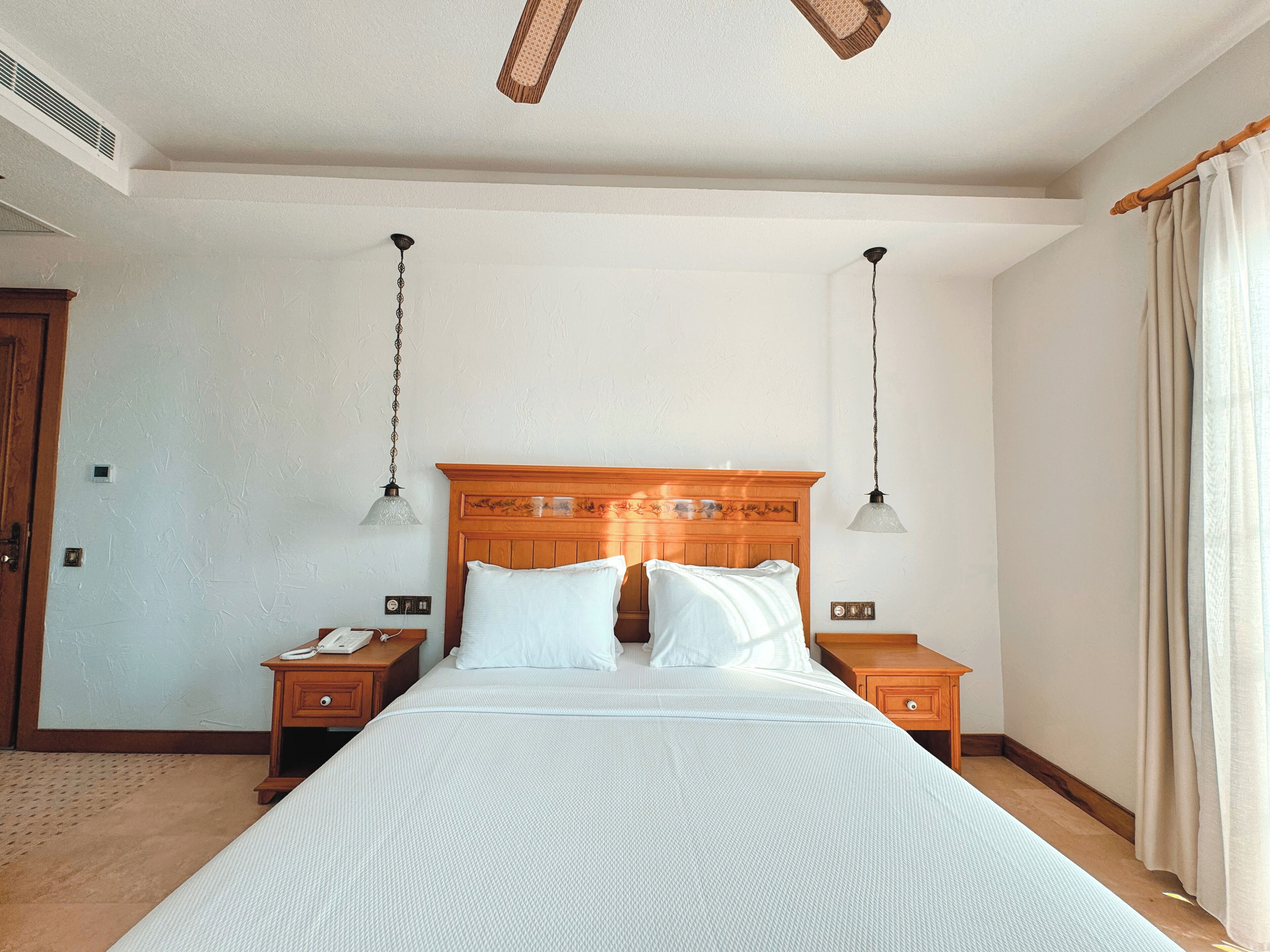 A neatly arranged hotel room with a large bed, two bedside tables, a wooden headboard, and two hanging lamps.