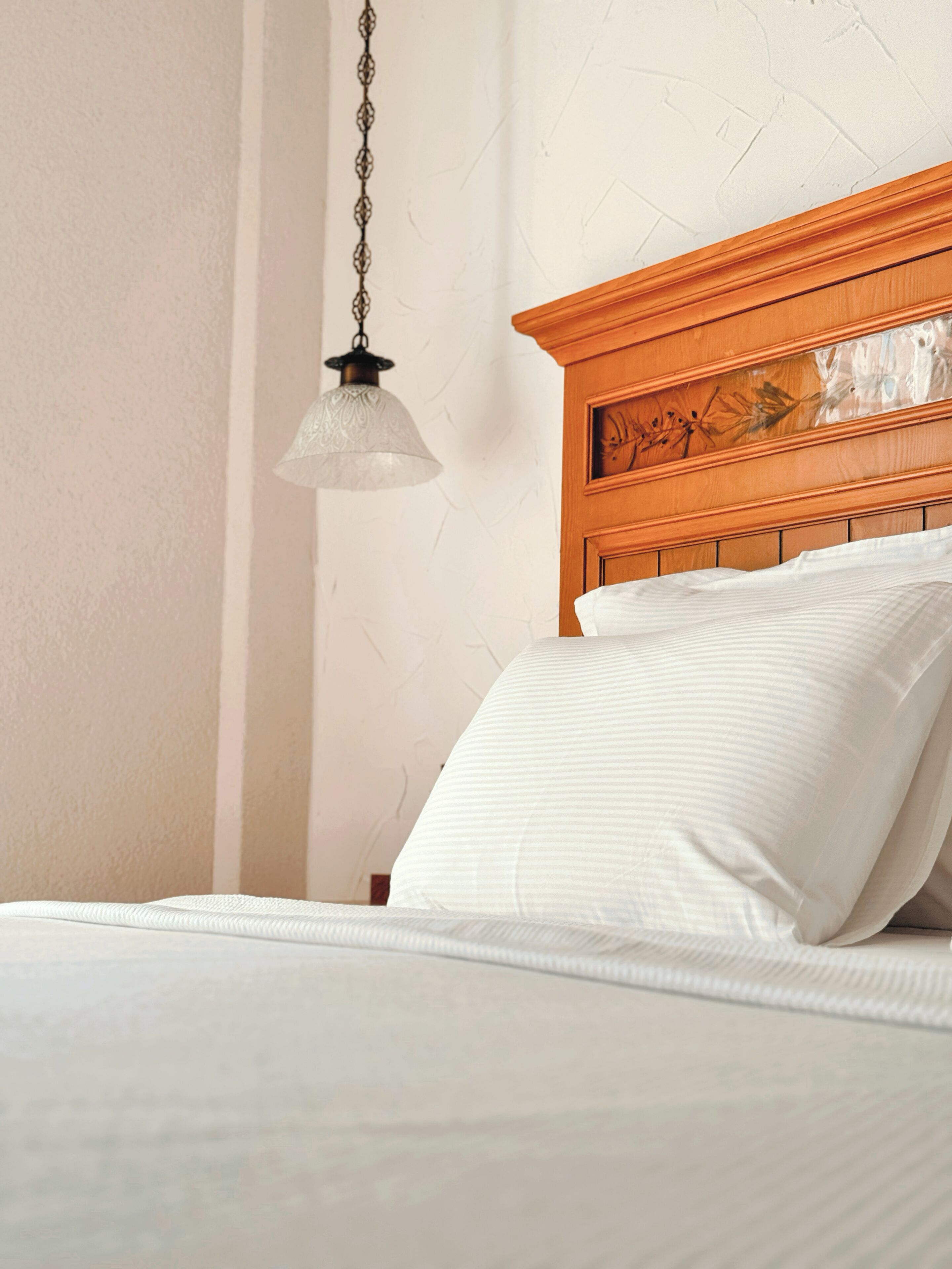 A neatly made bed with white linens and a wooden headboard, a hanging light fixture, and a plain wall.