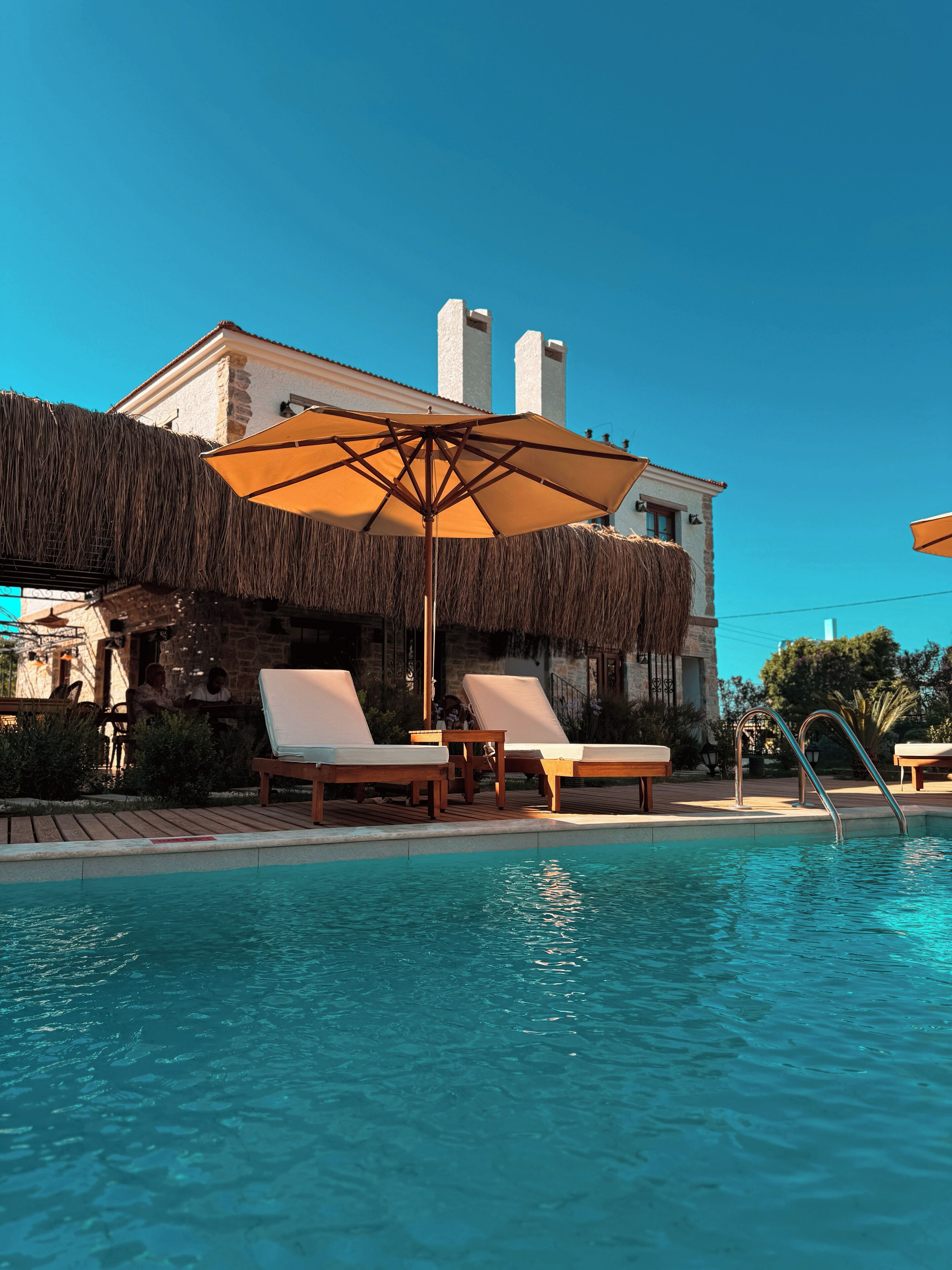 A pool area with lounge chairs and an umbrella, a building with a stone facade, and a clear blue sky.