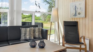A modern living room with a black leather sofa, a wooden coffee table, and a black leather chair. There are two decorative glass vases on the table. A large window offers a view of the outdoors, and a painting is on the wall.