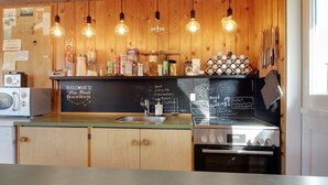 Una cocina con armarios de madera, un lavamanos de acero inoxidable, un microondas y una estufa. Hay bombillas colgantes y un menú en una pizarra en la pared.