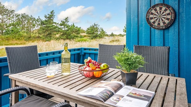 Una mesa de madera con una botella, vasos, una cesta de frutas, una revista y un dardo, todo sobre un fondo azul.