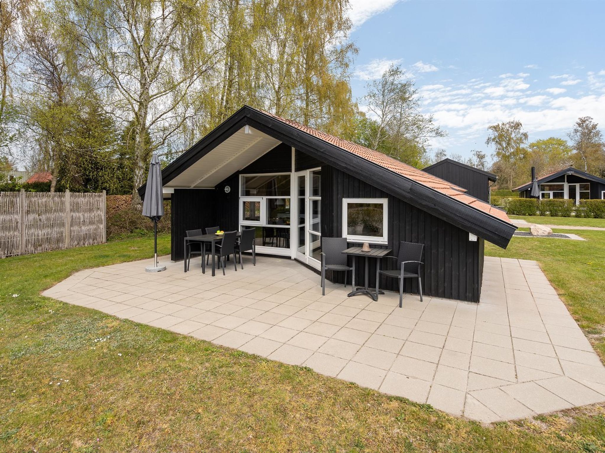 A modern house with a patio area featuring a table and chairs, surrounded by a fenced yard and trees.