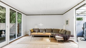 A modern living room with a wooden floor, a large sofa, a coffee table, and a dining table. There is a sliding glass door leading to an outdoor area. - "Malti" - 300m from the sea by Interhome (Sæby)