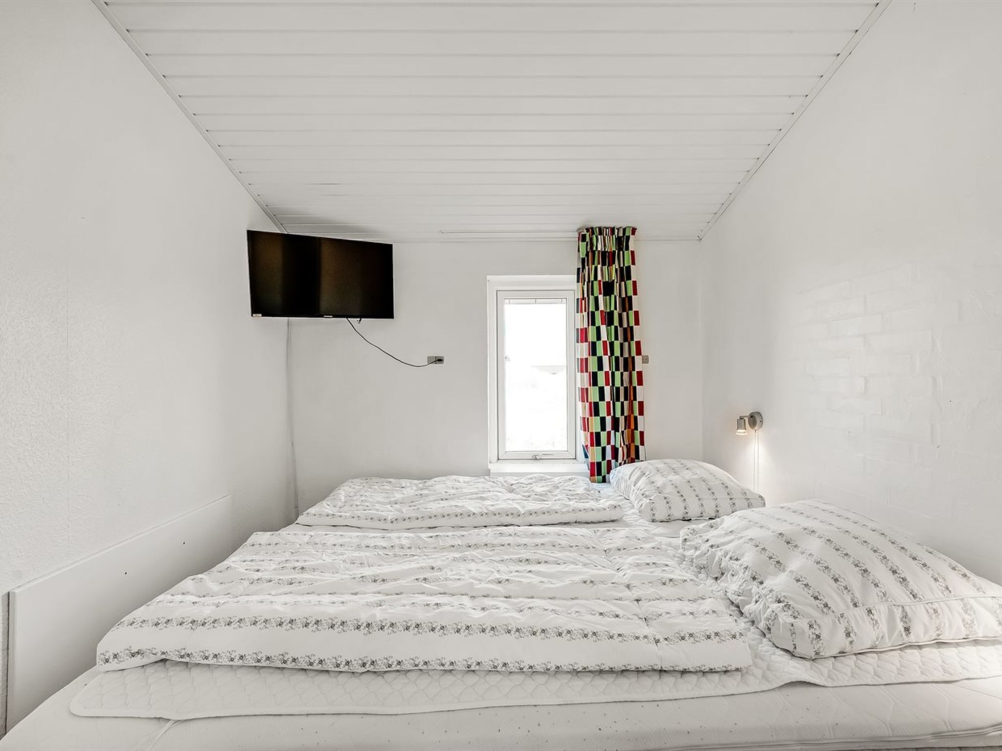 Een slaapkamer met een bed, een aan de muur bevestigde televisie, een raam met gordijn en een muur met baksteenmotief.