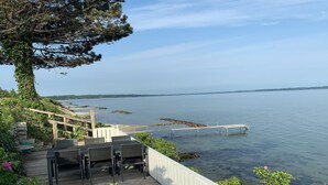 Une terrasse en bois avec des sièges extérieurs donnant sur une étendue d’eau calme, une jetée en bois s’avançant dans l’eau et un arbre aux feuilles vertes sur la gauche.