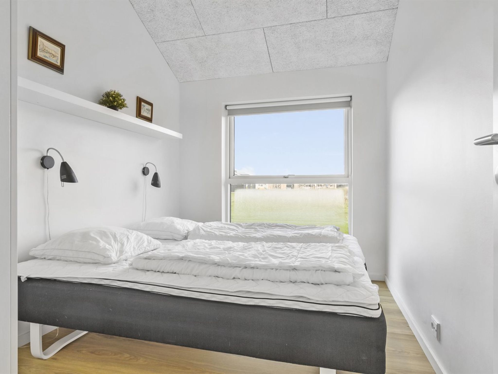 A modern bedroom with a bed, two wall-mounted lamps, a window with a view of a green field, and a small plant on a shelf.