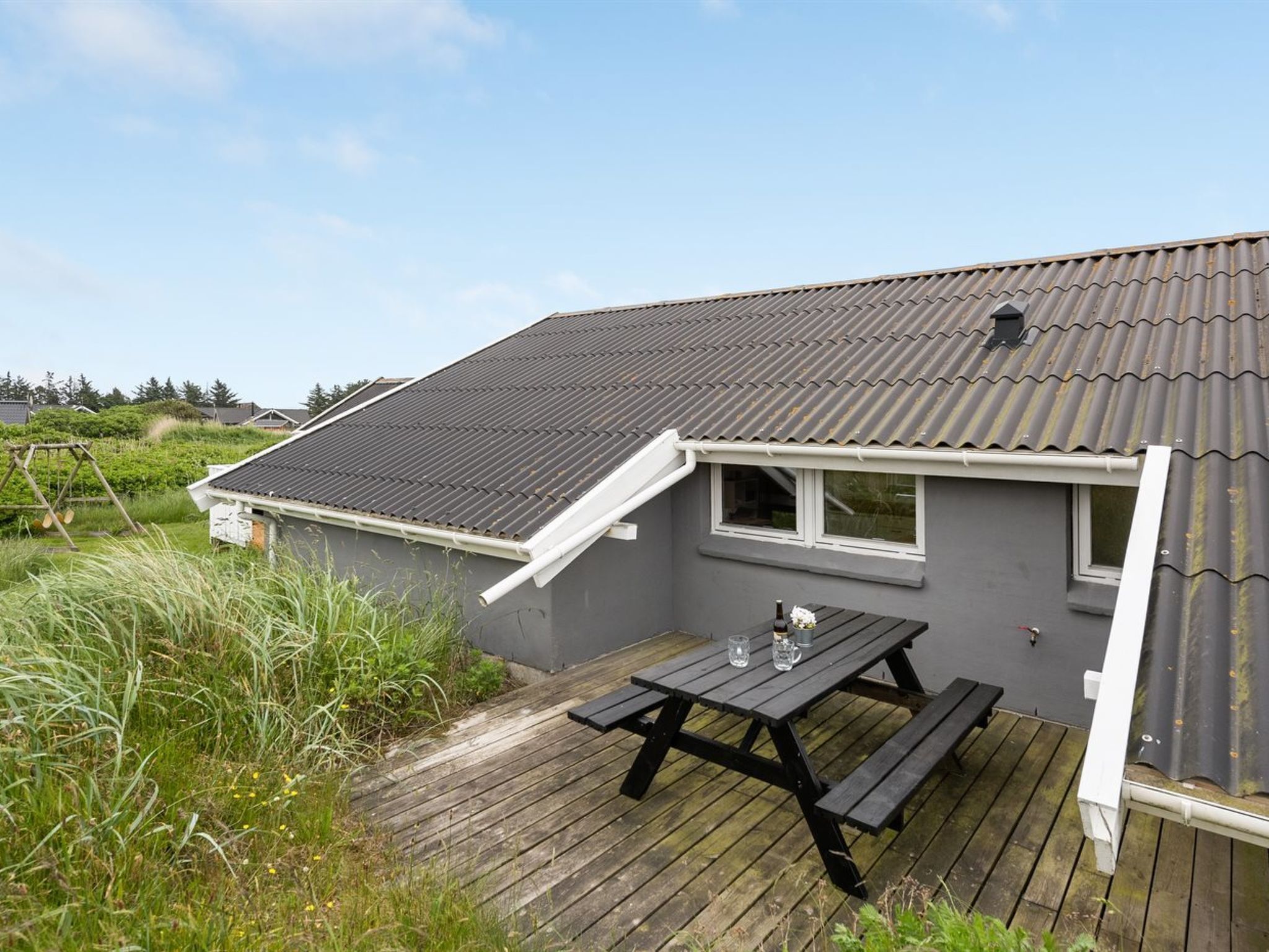 Une maison à l’extérieur gris, avec un toit en pente et une terrasse en bois équipée d’une table de pique-nique.