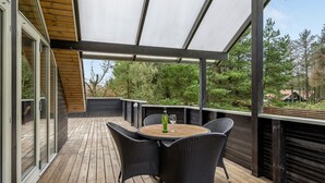 Un patio exterior con terraza de madera, un área de estar cubierta con sillas de mimbre y una mesa con una botella y un vaso.