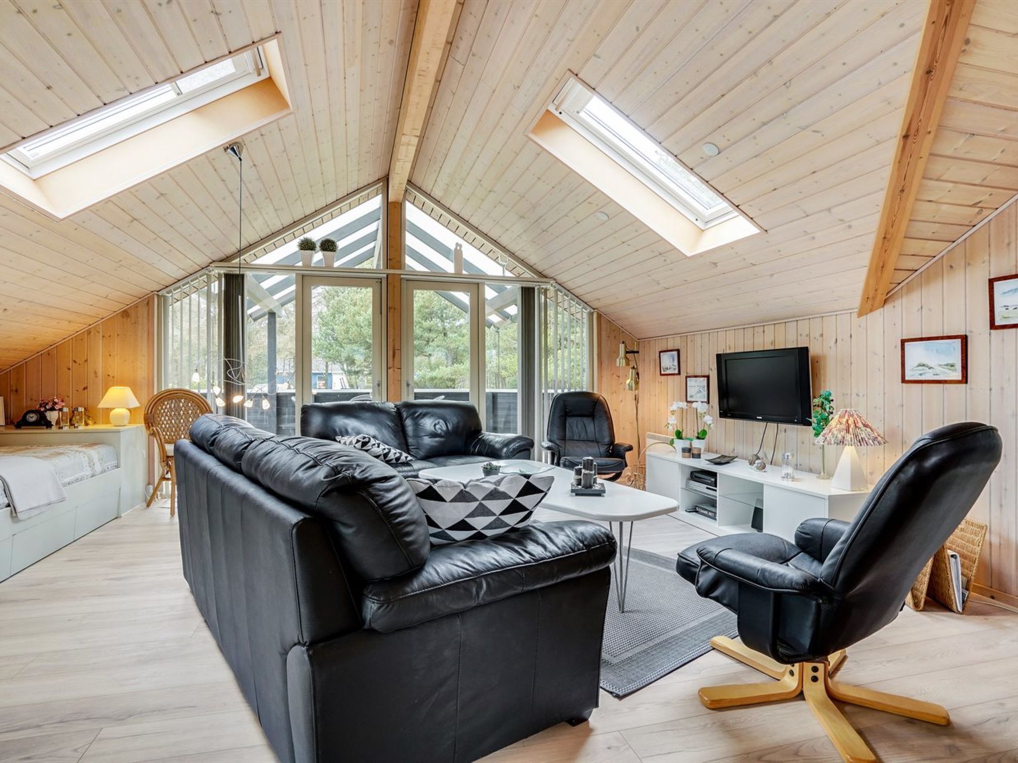 A modern loft-style room with a high ceiling, wooden beams, and skylights. It features a black leather sofa, a dining table, and a TV mounted on the wall.