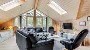 Una habitación moderna estilo loft con techo alto, vigas de madera y tragaluces. Cuenta con un sofá de cuero negro, una mesa de comedor y un televisor montado en la pared.