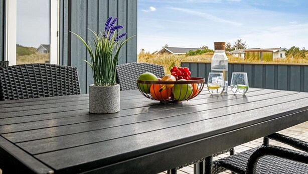 A wooden table with a bowl of fruit, a bottle of water, and a glass on a patio with wicker furniture and a potted plant. - "Estella" - 500m to the fjord by Interhome (Hvide Sande)
