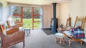 Ein Zimmer mit großem Fenster, Kaminofen, Korbmöbeln und Blick auf Grünflächen.