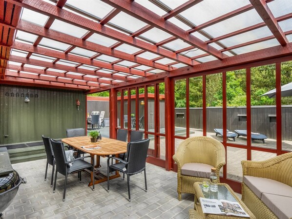 A covered patio with a wooden table and chairs, a grill, and wicker furniture.