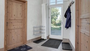 Een gang met een houten deur, een glazen deur naar buiten, een kapstok met kleding en een klein wandrek.