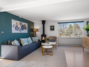 En opholdsstue med sofa, bord, lampe, pejs og udsigt gennem store vinduer.