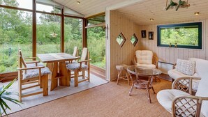 A cozy room with a wooden ceiling, large windows offering a view of greenery, a dining table with chairs, a sofa, and a small table with a glass top.