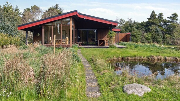 A modern house with a red roof, large glass windows, and a pond in the foreground.