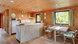 A wooden cabin with a kitchen and dining area, featuring a central island, wooden flooring, and large windows.