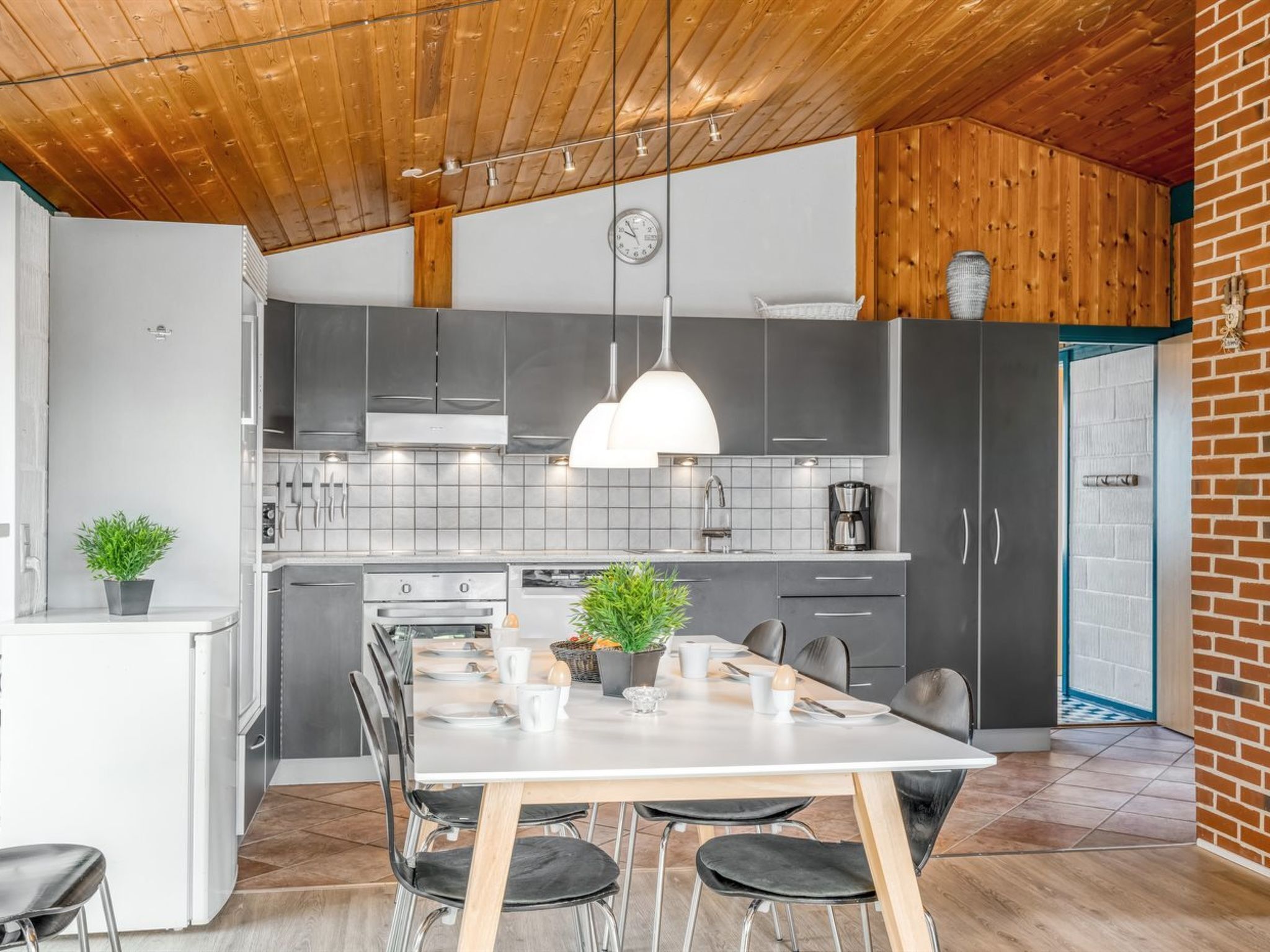 Een moderne keuken met grijze kastjes, een wit aanrecht en een houten plafond. Er staat een eettafel met stoelen en een kamerplant.