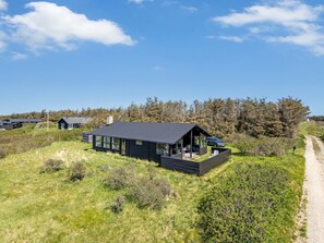 Une maison moderne aux couleurs sombres, dotée d’une grande terrasse et entourée de verdure, sous un ciel dégagé.