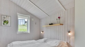 A bedroom with a bed, a window, a wall-mounted shelf with flowers, and a framed picture.
