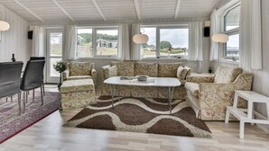 A spacious living room with a white ceiling, large windows, a central white coffee table, floral-patterned sofas, a dining table with chairs, and a patterned rug.