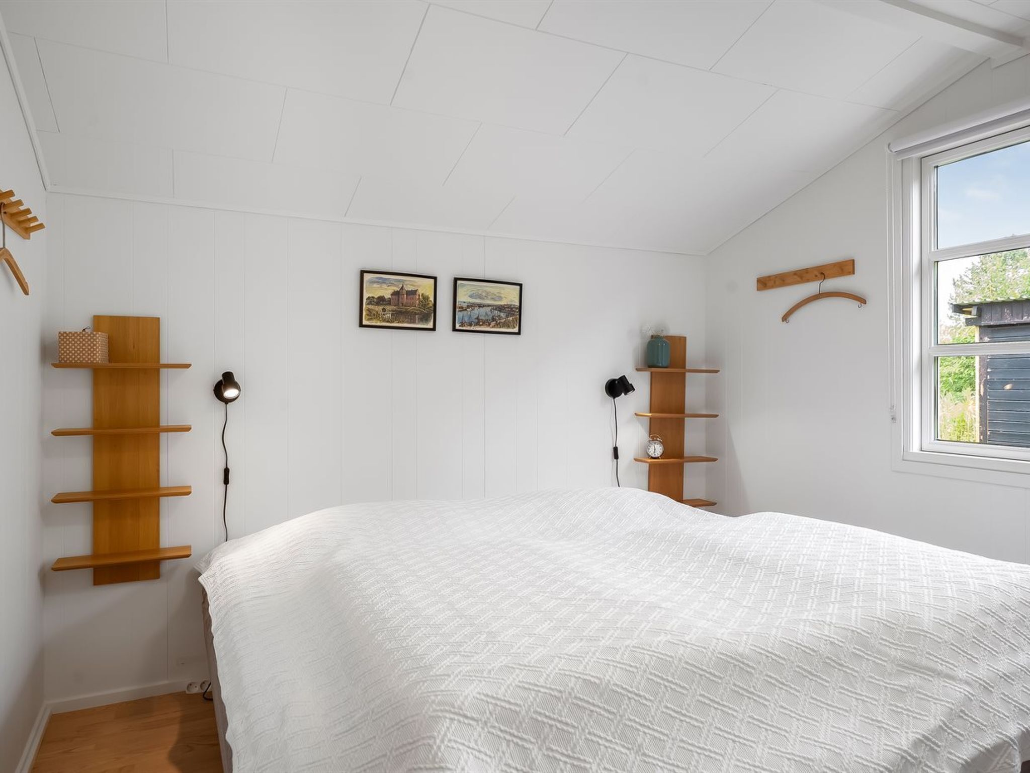 Une chambre à coucher avec un lit, des étagères en bois, deux tableaux encadrés, une fenêtre et un porte-manteau suspendu.