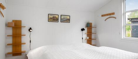 Une chambre à coucher avec un lit, des étagères en bois, deux tableaux encadrés, une fenêtre et un porte-manteau suspendu.
