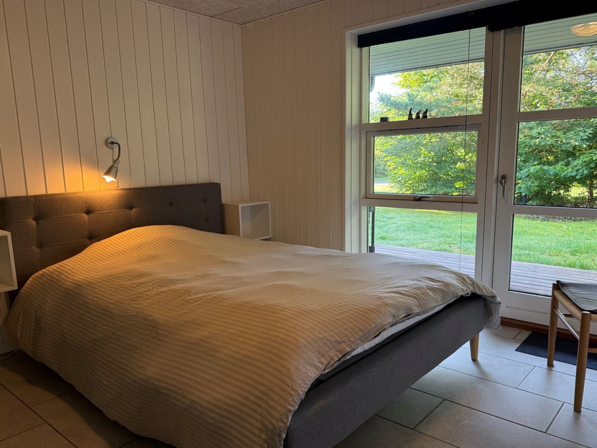 A bedroom with a bed, a nightstand, a wall-mounted lamp, and a sliding glass door leading to a garden.