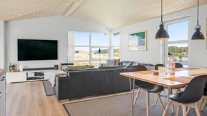 A modern living room with a large black TV, a grey sofa, a wooden dining table with black chairs, and large windows offering a view of the outdoors. - "Ingela" - 350m from the sea by Interhome (Løkken)