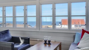 Ein Zimmer mit Blick auf Strand und Meer durch große Fenster. Ein Holztisch mit Gläsern und ein Stuhl sind vorhanden.