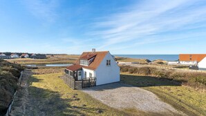 Ein Küstenhaus mit weißer Fassade und rotem Dach, umgeben von einem Schotterweg und einer Rasenfläche, mit Blick auf das Meer und weitere Häuser im Hintergrund.