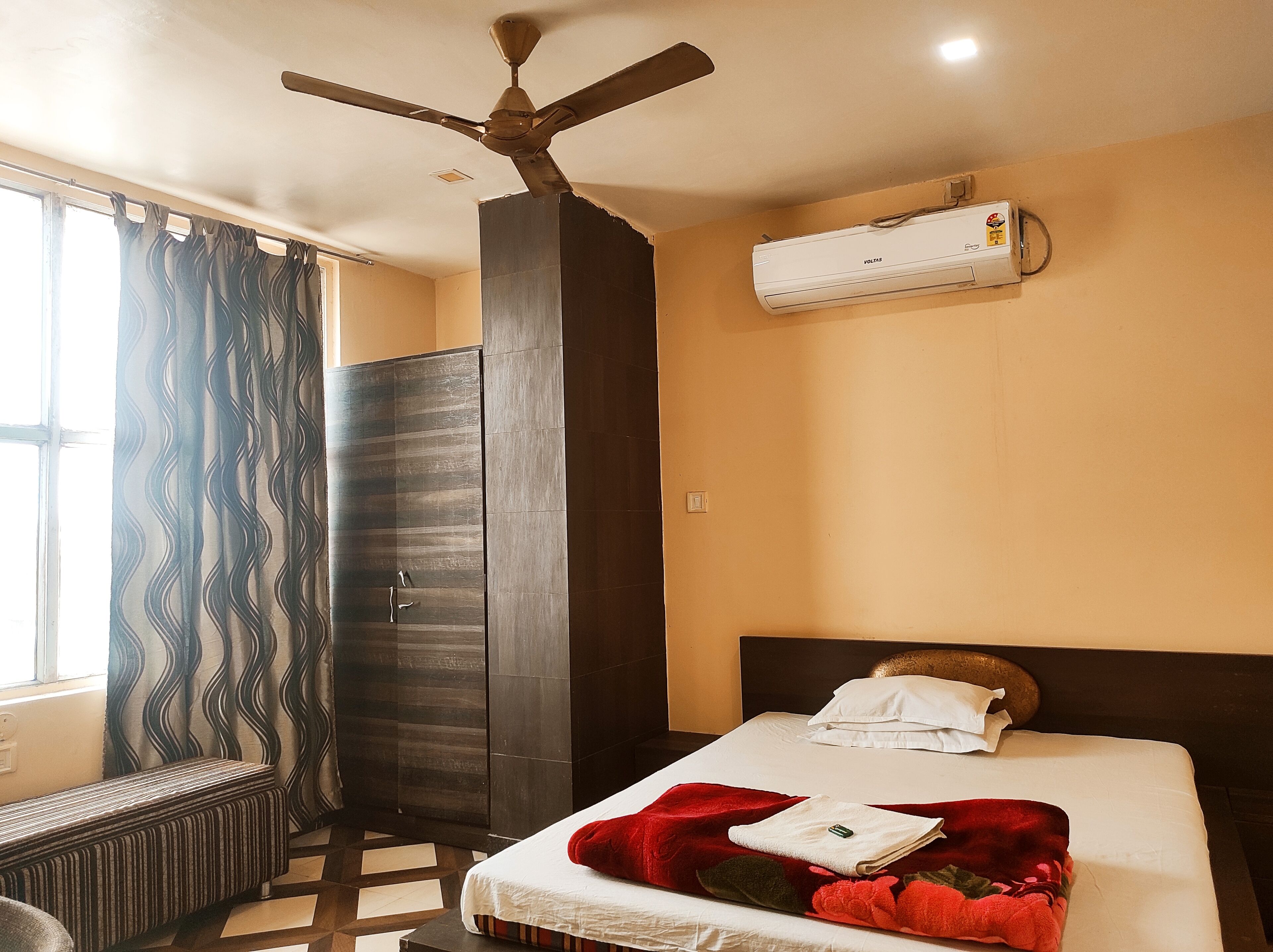 A hotel room with a bed, a ceiling fan, an air conditioning unit, a wardrobe, and a window with curtains.