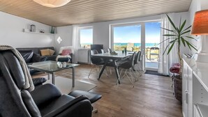 Un saló modern amb una taula de café de vidre, un sofà de pell negra i una zona de menjador amb taula i cadires blanques. Grans finestres ofereixen una vista de la platja.